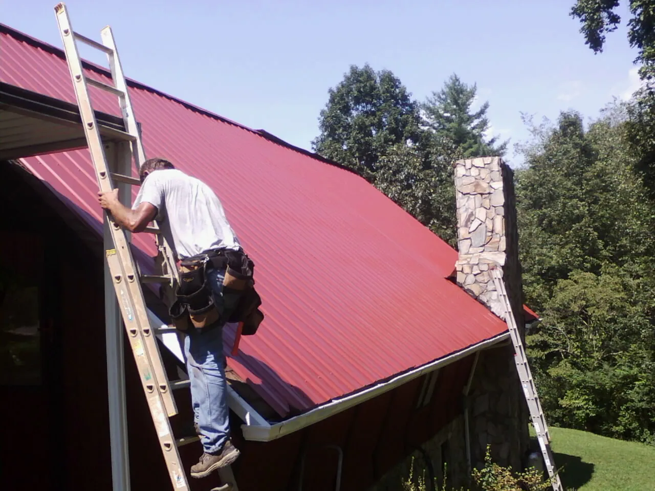 Roofer on a ladder beside a red metal roof and stone chimney — Soffit & Fascia Repair by Roofing Brothers in Pentwater